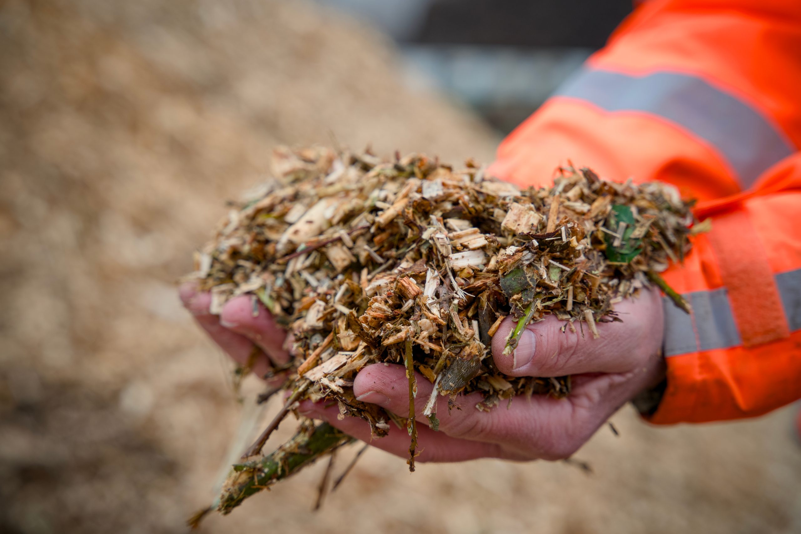 A person wearing an orange safety jacket holds a handful of wood chips and plant material, showcasing a mix of small twigs, leaves, and shredded wood. In the background, a pile of similar material is visible