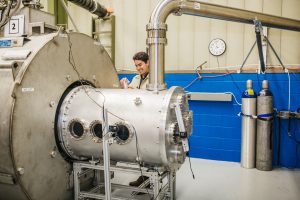 A man stands next to a large, metallic chamber with multiple viewing ports and connected pipes. He is holding a small object and appears to be examining the chamber, which is part of a laboratory setting. In the background, there are gas cylinders and a clock on the wall