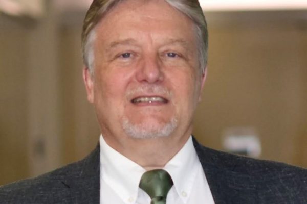 A man with short gray hair and a goatee smiles at the camera. He is wearing a dark suit jacket over a white shirt and a green tie. The background is blurred, suggesting an indoor setting