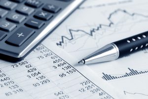 A calculator, a silver pen, and printed financial documents with numerical data and graphs are arranged on a surface. The documents display various figures and charts related to financial analysis