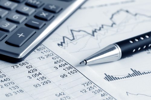 A calculator, a silver pen, and printed financial documents with numerical data and graphs are arranged on a surface. The documents display various figures and charts related to financial analysis