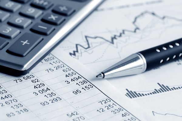 A calculator, a silver pen, and printed financial documents with numerical data and graphs are arranged on a surface. The documents display various figures and charts related to financial analysis