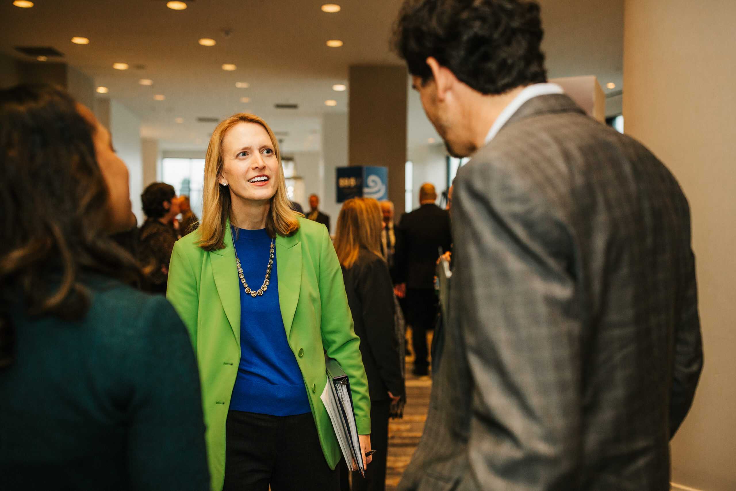 A woman with long blonde hair, wearing a bright green blazer and a blue top, is engaged in conversation with a man in a gray plaid suit. They are in a crowded indoor setting, likely at a conference or networking event, with other attendees visible in the background