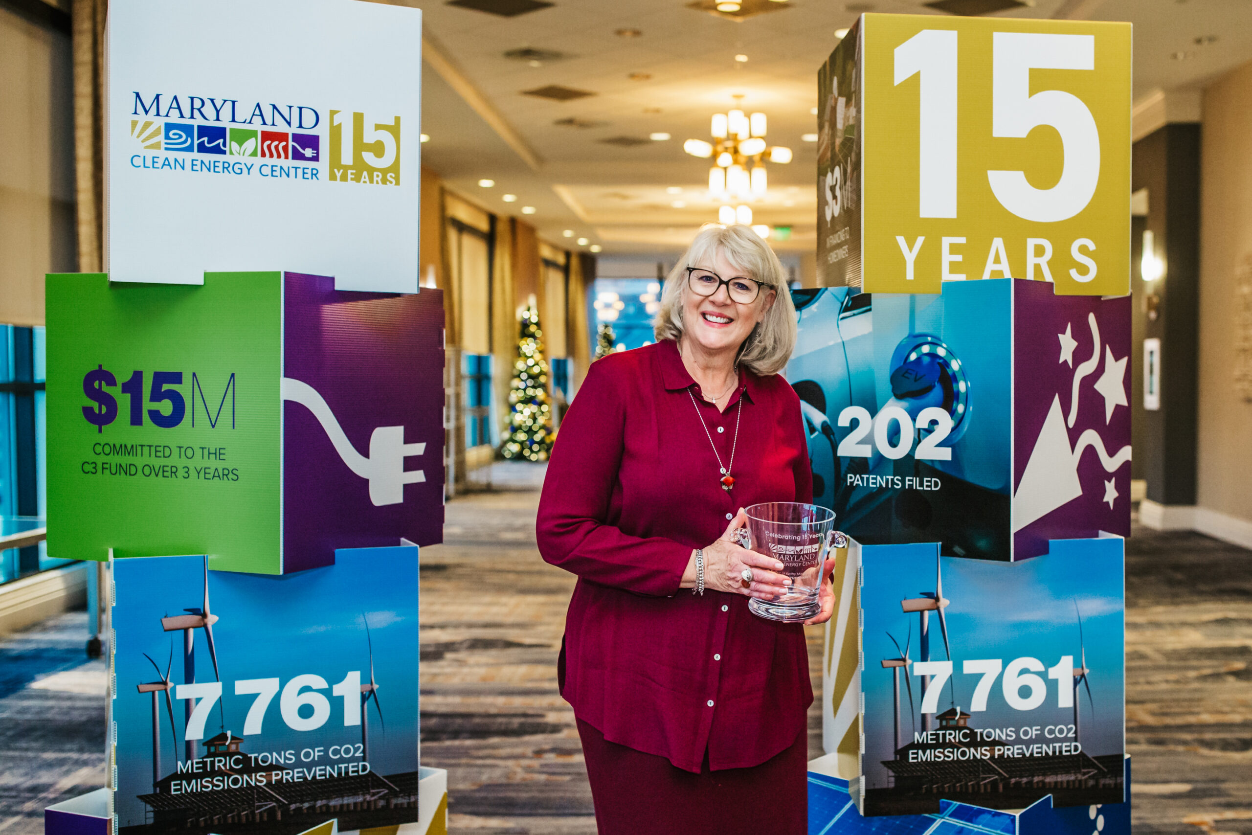 A woman with blonde hair and glasses stands holding a clear award in front of two large display boards. The boards highlight the Maryland Clean Energy Center's 15-year anniversary.