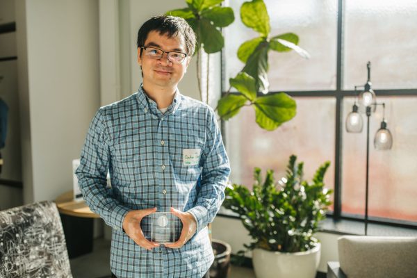 A man with glasses and short black hair stands in a well-lit room, holding a clear award or trophy. He is wearing a blue and white checkered shirt and has a name tag on his left side. In the background, there are green plants and a window with natural light