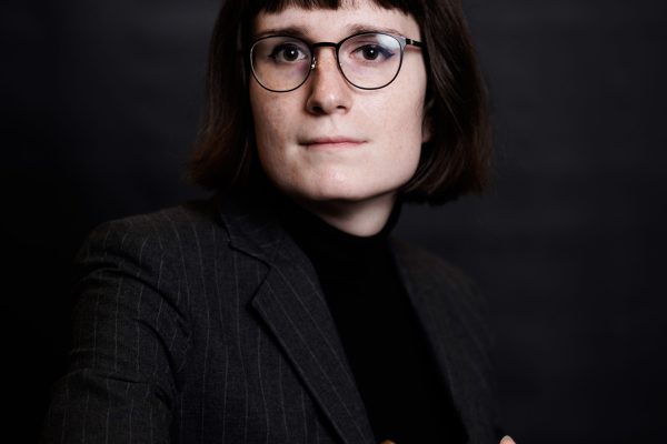 A person with short brown hair and glasses is wearing a dark blazer over a black turtleneck. They are seated, looking directly at the camera with a serious expression, and have a gold bracelet on their wrist. The background is dark and neutral