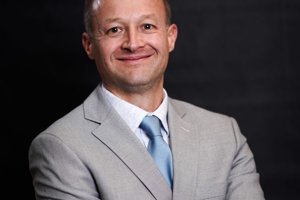 A man with short light brown hair smiles while wearing a light gray suit and a blue tie. He stands with his arms crossed against a dark background