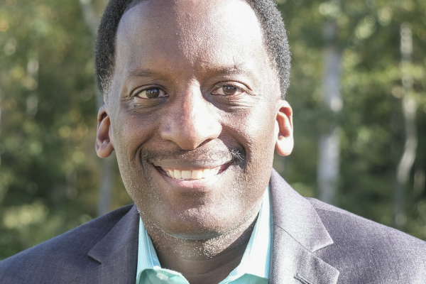 A man with short, curly black hair smiles while wearing a light blue shirt and a dark blazer. He is outdoors, with trees in the background