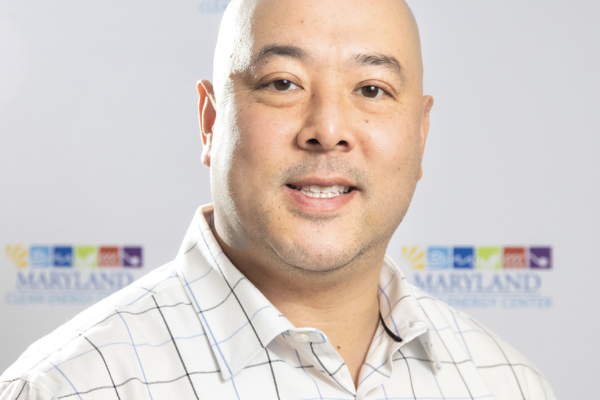 A man with a shaved head and a slight smile is wearing a checkered white shirt. He is standing in front of a light-colored background featuring a logo that includes the word "MARYLAND" and colorful squares