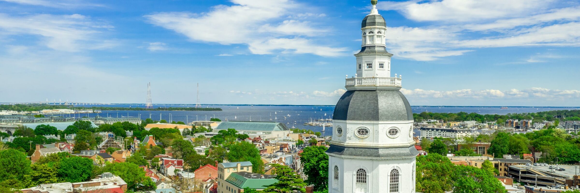 A view of a city with a prominent dome structure. The dome is part of a historic building, likely a government or civic structure. In the background, there is a body of water with boats and a clear blue sky.