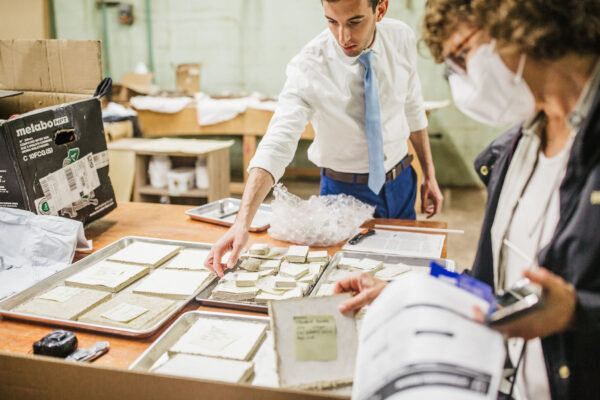 A person is sorting through small packets on a tray, while another individual holds a document and a phone. The setting appears to be an office or workspace with various materials and equipment visible in the background
