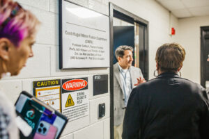 A group of people stands in a hallway outside a door labeled "Centrifugal Mirror Fusion Experiment (CMFX)" with a sign indicating the professor's name and affiliation. One person, a man in a blazer, is speaking to the group, while another person, a woman with pink hair, holds a tablet. A caution sign is visible on the wall, warning of a strong magnetic field