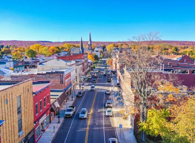A view of a small town street lined with shops and buildings, featuring a mix of red and brick facades. In the background, two church steeples rise above the rooftops, and colorful autumn trees are visible on the hills beyond. Cars are parked along the street and driving through the town. The sky is clear and blue
