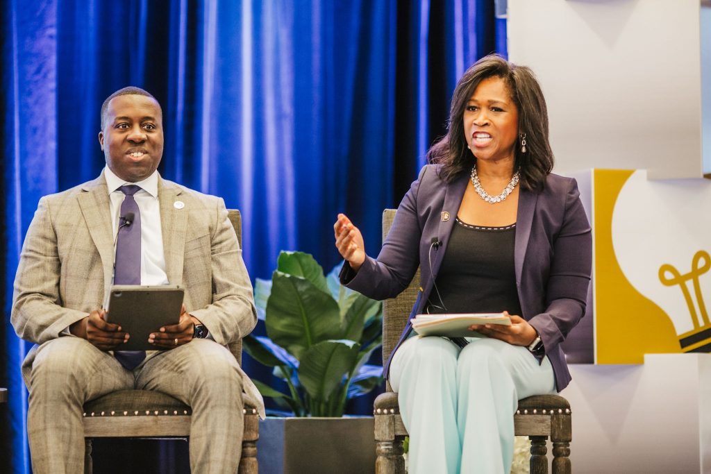 A man in a gray suit and a woman in a black blazer and light blue pants are seated on stage during a panel discussion. The man holds a tablet and looks attentively at the woman, who is gesturing with her hand while speaking. The background features blue drapes and a decorative light bulb graphic
