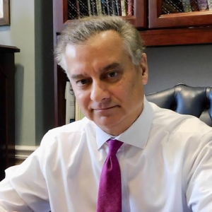 A man with short, light brown hair and a slight smile sits at a desk, wearing a white dress shirt and a pink tie. Behind him are wooden shelves filled with books