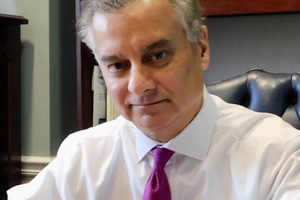 A man with short, light brown hair and a slight smile sits at a desk, wearing a white dress shirt and a pink tie. Behind him are wooden shelves filled with books