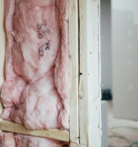 Insulation material made of pink fiberglass is installed between wooden framing in a wall. The surrounding area shows unfinished drywall and construction debris on the floor