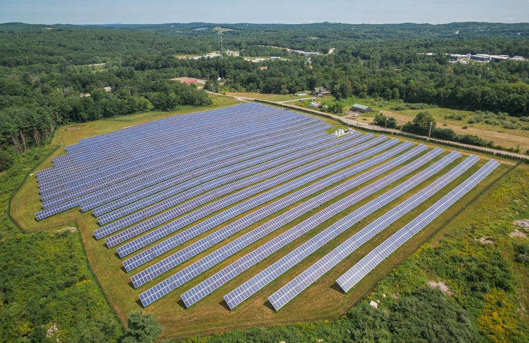 A large solar farm featuring numerous rows of solar panels arranged in a grid pattern, set in a green landscape with trees and hills in the background. The panels cover a significant area of land, reflecting sunlight