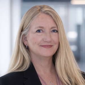 A woman with long, straight blonde hair and blue eyes smiles confidently. She is wearing a black blazer over a light pink blouse and is positioned in an office setting with blurred background elements