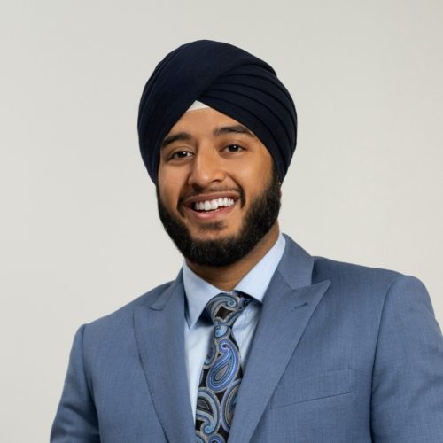 A man wearing a dark blue turban and a light blue suit smiles at the camera. He has a well-groomed beard and is wearing a patterned tie. The background is plain and light-colored