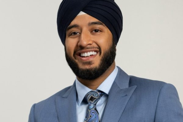 A man wearing a dark blue turban and a light blue suit smiles at the camera. He has a well-groomed beard and is wearing a patterned tie. The background is plain and light-colored