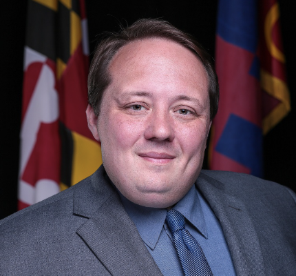 A man with short brown hair and a slight smile is wearing a gray suit and a blue shirt with a tie. He is positioned in front of a dark background featuring Maryland state flags