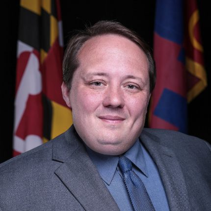 A man with short brown hair and a slight smile is wearing a gray suit and a blue shirt with a tie. He is positioned in front of a dark background featuring Maryland state flags