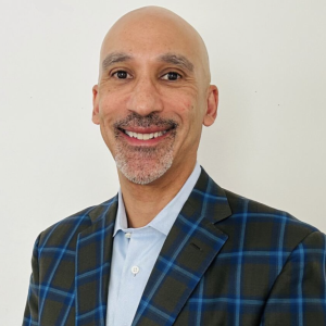 A man with a bald head and a neatly trimmed beard smiles at the camera. He is wearing a blue and green plaid blazer over a light blue shirt. The background is plain and white