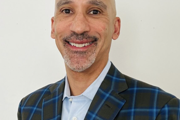 A man with a bald head and a neatly trimmed beard smiles at the camera. He is wearing a blue and green plaid blazer over a light blue shirt. The background is plain and white