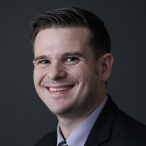 A man with short, styled hair and a friendly smile is wearing a dark suit and a light-colored shirt. He is facing slightly to the right against a dark background