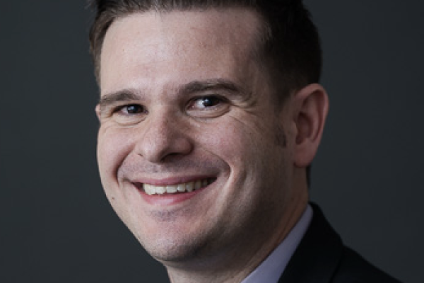 A man with short, styled hair and a friendly smile is wearing a dark suit and a light-colored shirt. He is facing slightly to the right against a dark background