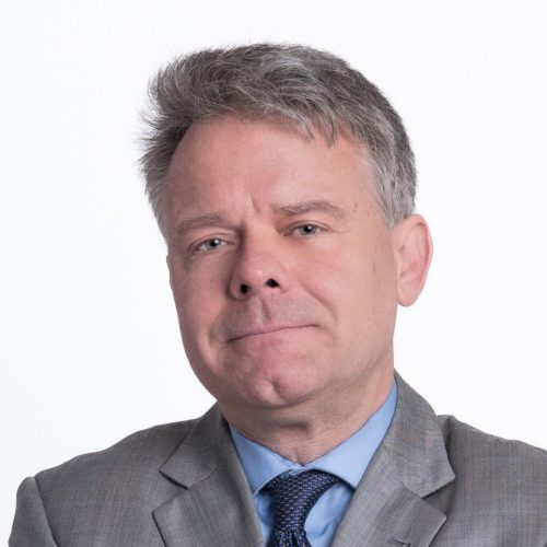 A man with short gray hair and a slight smile is wearing a light gray suit and a blue dress shirt with a tie. He is facing the camera against a plain white background