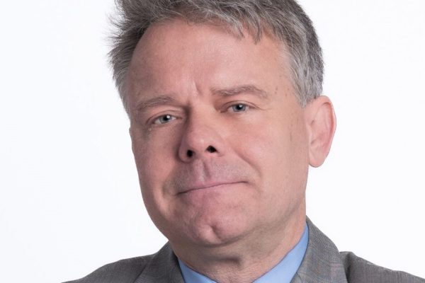 A man with short gray hair and a slight smile is wearing a light gray suit and a blue dress shirt with a tie. He is facing the camera against a plain white background