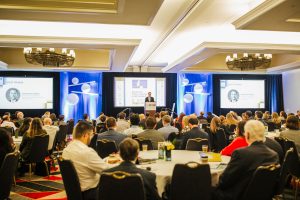 A speaker stands at a podium in front of a large audience in a conference room. Two large screens display information and a presentation. Attendees are seated at round tables, some taking notes or looking at the screens. The room is well-lit with decorative lighting and a modern design