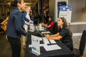 A woman in a black dress sits at a registration table, smiling and looking at another woman who is standing and speaking to her. The table has signs indicating check-in and sections labeled "Q-Z" and "L-P." Two other individuals are visible in the background, one seated and one standing. The setting appears to be a conference or event