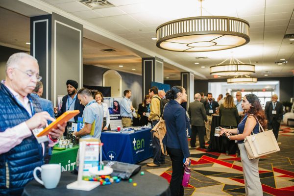 A busy conference setting with attendees engaging in conversations and networking. Several tables display promotional materials and products.