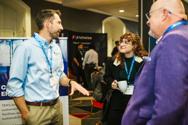 A man in a light blue shirt speaks to two people at a conference. One person, a woman with curly hair, holds a coffee cup and listens attentively. The other person, a man wearing glasses and a purple jacket, appears engaged in the conversation. A banner in the background displays the logo for TEN