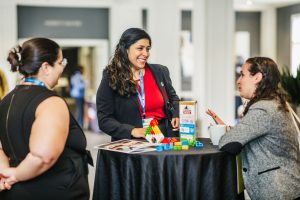 Three women are engaged in conversation around a small table. One woman, wearing a black blazer and red shirt, is smiling and holding colorful building blocks. The other two women, one in a black dress and the other in a gray blazer, are listening attentively. The setting appears to be a conference or networking event, with people visible in the background