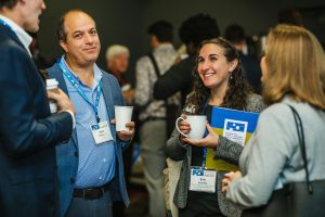 A group of four people engage in conversation at a conference. Two individuals, a man with short hair and a woman with curly hair, are smiling and holding coffee cups. The man is wearing a blue shirt and a blazer, while the woman is holding a folder and wearing a gray sweater. Two other people are partially visible in the background, also participating in the discussion