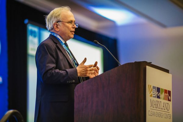 A man in a suit stands at a podium, speaking to an audience. He gestures with his hands while addressing attendees at a conference, with a backdrop featuring the Maryland Clean Energy Center logo