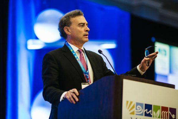 A man in a black suit and pink tie stands at a podium, gesturing with his right hand while holding glasses in his left. He appears to be speaking at a conference, with a blue and white backdrop featuring abstract shapes. A name tag is visible around his neck