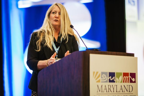 A woman with long blonde hair stands at a podium, speaking to an audience. She is wearing a black blazer and appears to be engaged in a presentation. The podium has a sign that reads "MARYLAND" along with various colorful symbols