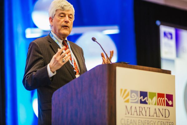 A man in a suit speaks at a podium with the Maryland Clean Energy Center logo. He gestures with his hands while addressing an audience at a conference. The background features blue lighting and banners related to the event