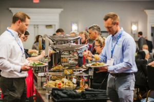 A group of people at a buffet-style meal, serving themselves from a food station. Several individuals are holding plates with food, while others are in conversation. The setting appears to be a conference or event, with tables and chairs in the background