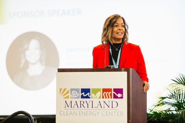 A woman with shoulder-length brown hair, wearing a red blazer, stands at a podium with a sign that reads "MARYLAND CLEAN ENERGY CENTER." She is speaking to an audience, with a blurred image of her in the background