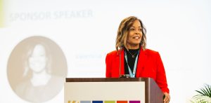 A woman with shoulder-length brown hair, wearing a red blazer, stands at a podium with a sign that reads "MARYLAND CLEAN ENERGY CENTER." She is speaking to an audience, with a blurred image of her in the background