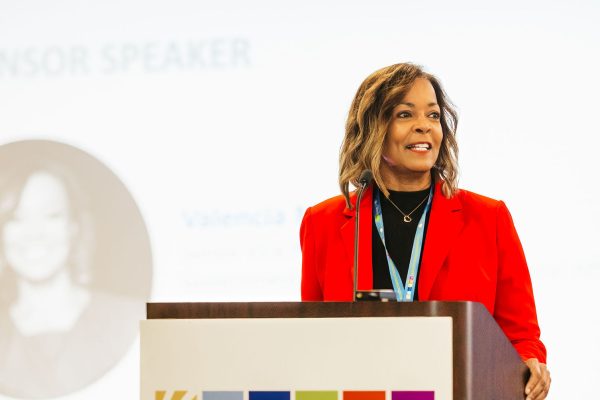 A woman with shoulder-length brown hair, wearing a red blazer, stands at a podium with a sign that reads "MARYLAND CLEAN ENERGY CENTER." She is speaking to an audience, with a blurred image of her in the background