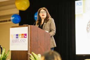 A woman with long brown hair and glasses stands at a podium, speaking to an audience. She is wearing a plaid blazer and appears to be engaged in a presentation. The background features colorful balloons and a screen displaying information related to the Maryland Clean Energy Center
