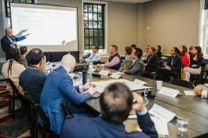 A group of people is seated around a conference table in a meeting room. A presenter stands at the front, pointing to a slide on a screen. Attendees are engaged, some taking notes, while others are looking at the presenter. The room has large windows allowing natural light to enter