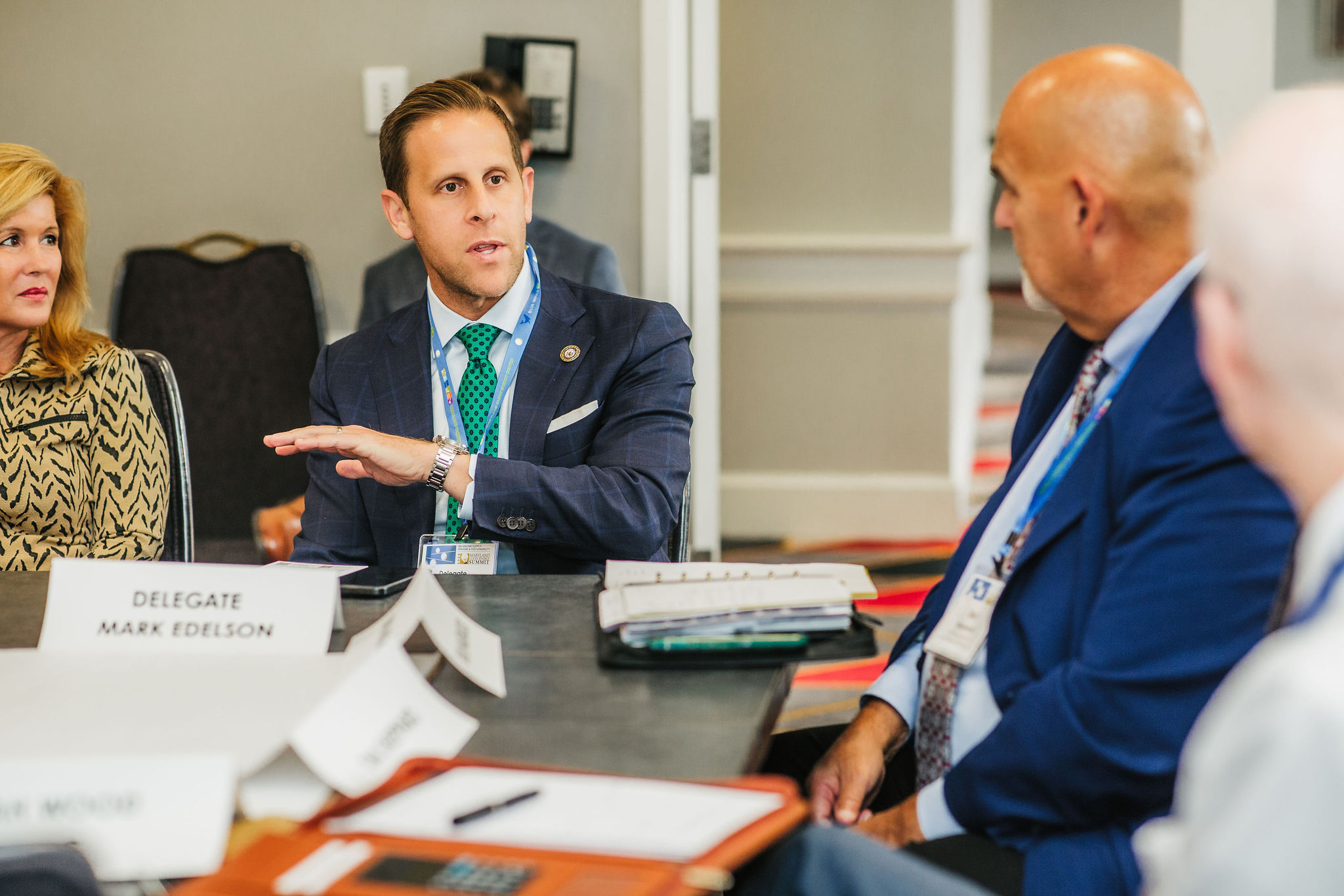 A man in a suit gestures while speaking at a roundtable discussion. He has short hair and is wearing a green tie. Two other individuals are seated nearby, one with blonde hair and the other bald, both listening attentively. Nameplates on the table indicate the presence of delegates